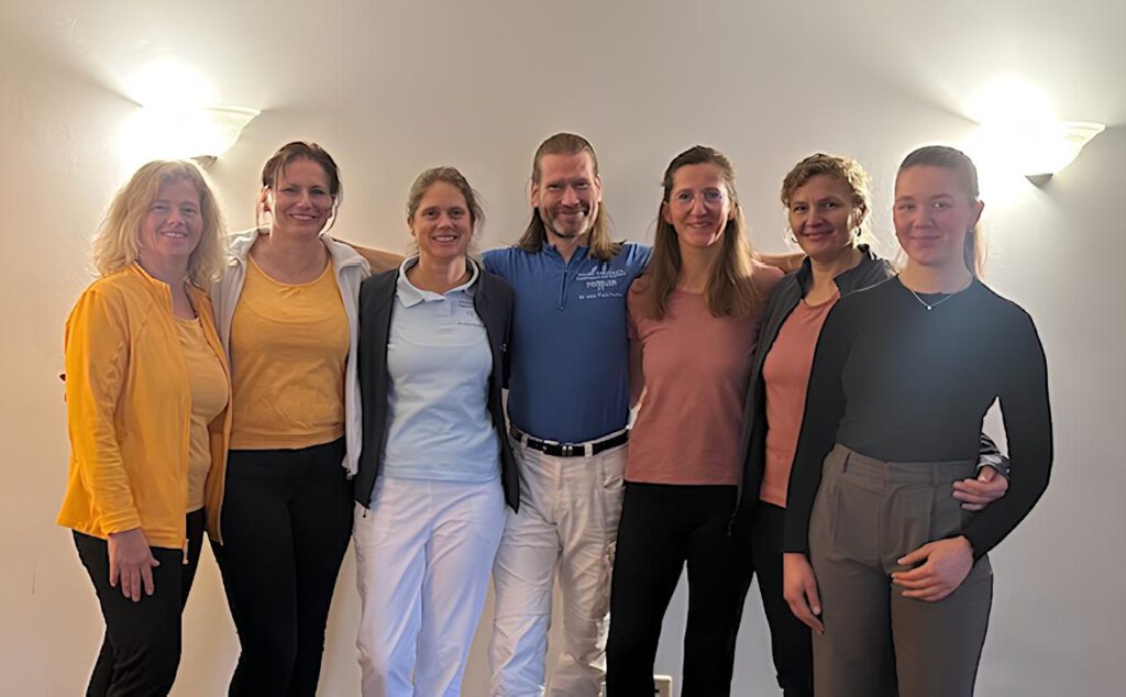 Sieben Erwachsene stehen in einem Haus zusammen und lächeln in die Kamera, während die Wand hinter ihnen warm beleuchtet ist.